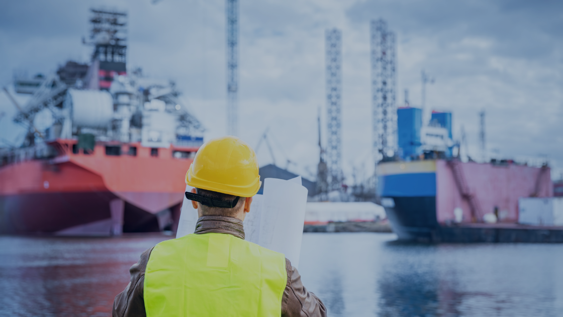 Ingenieur mit Schutzhelm betrachtet Baupläne vor Schiffen im Hafen, Symbol für Rohrfertigung und alternative Produktionsprozesse im Schiffbau wie Rohrbiegen statt Schweißen.